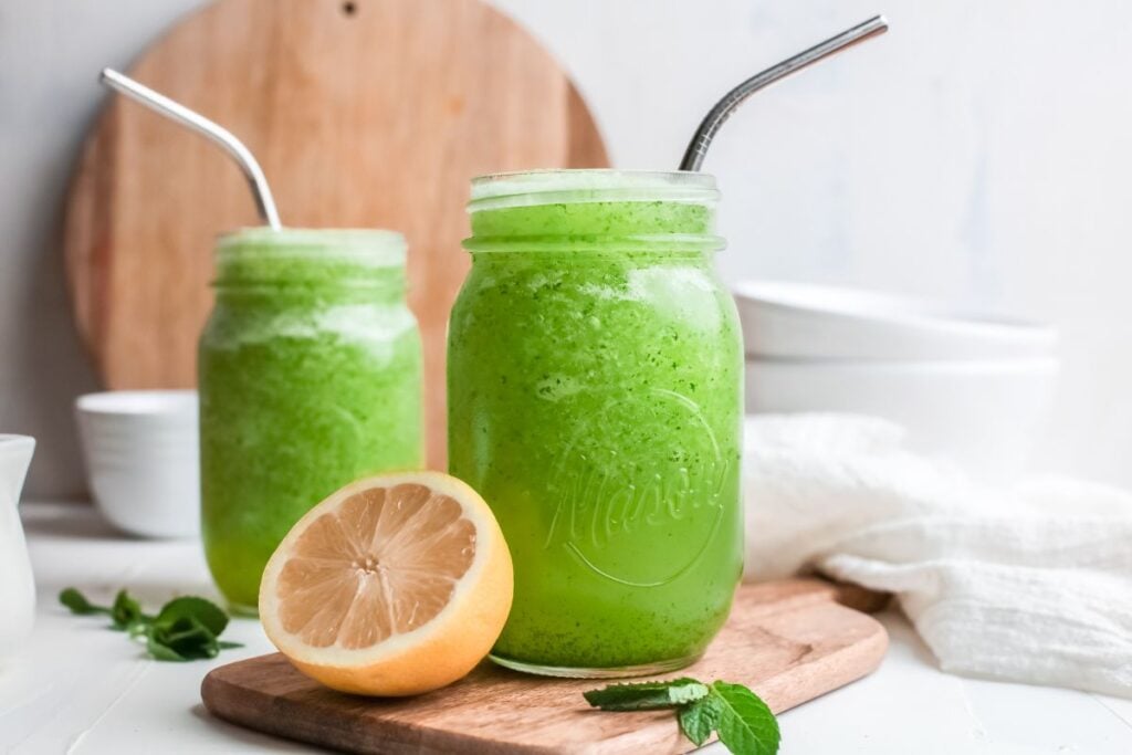 Two mason jars filled with a refreshing Limonana—an invigorating lemon and mint drink—sit with metal straws, a lemon half, and fresh mint leaves on a wooden board.