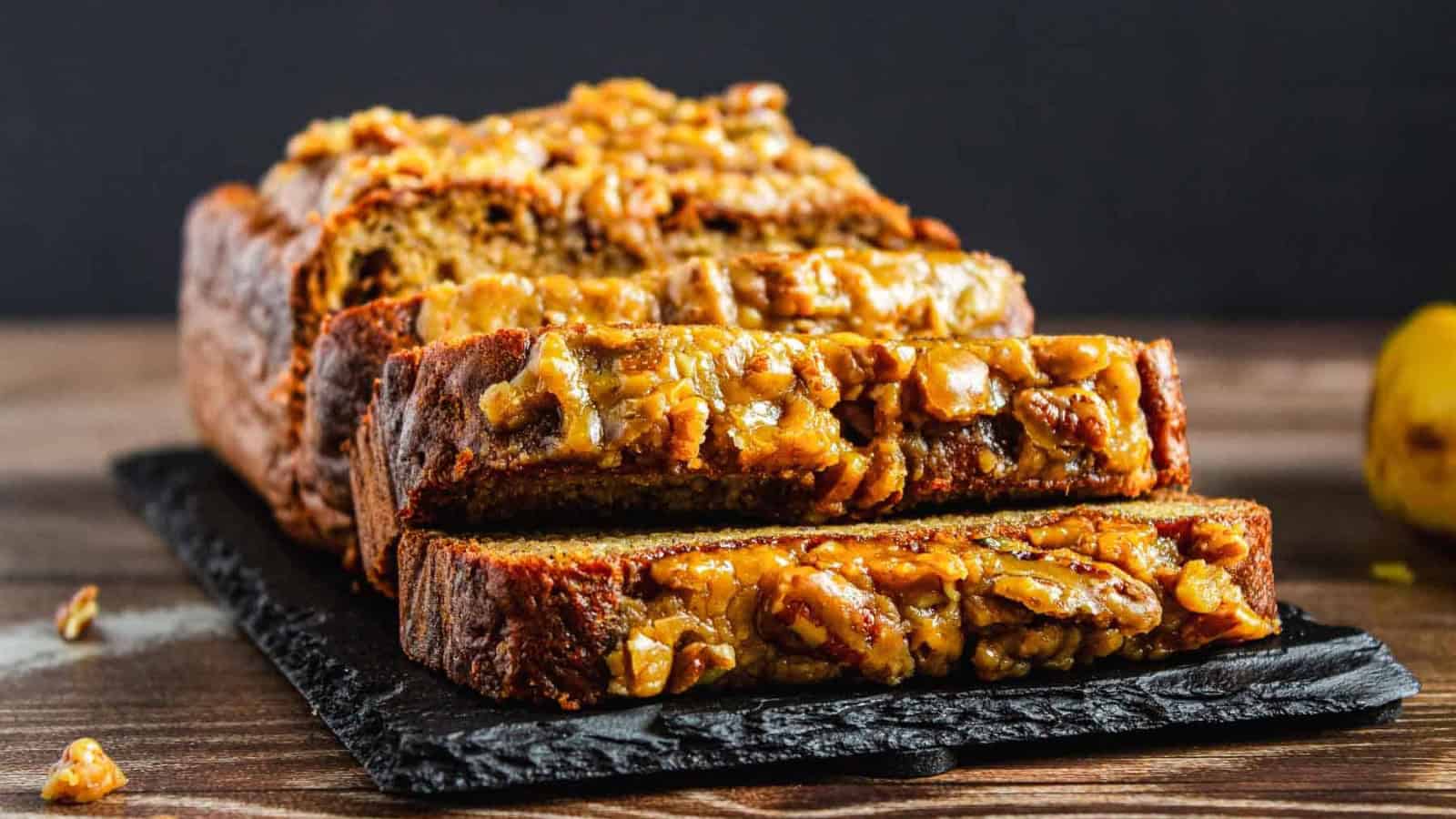 Sliced banana bread loaf topped with walnuts, displayed on a black slate board on a wooden table.