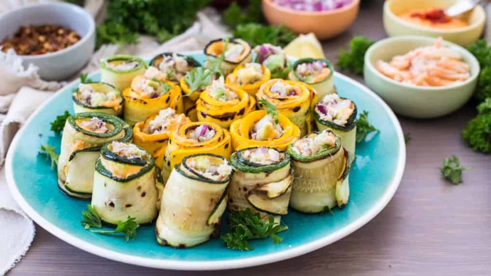 A blue plate with neatly arranged zucchini and yellow squash roll-ups filled with diced vegetables and herbs. Small bowls of ingredients and fresh parsley are visible in the background.