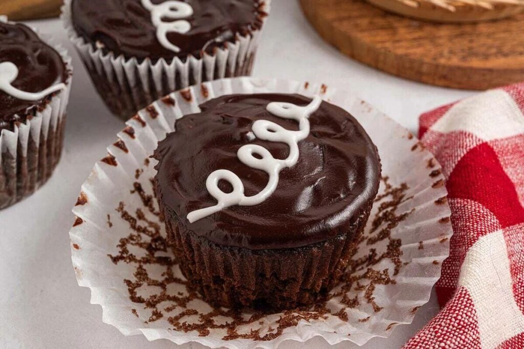Homemade Hostess Cupcakes: Chocolate cupcake with glossy frosting and a white icing swirl, sitting on an opened cupcake wrapper.