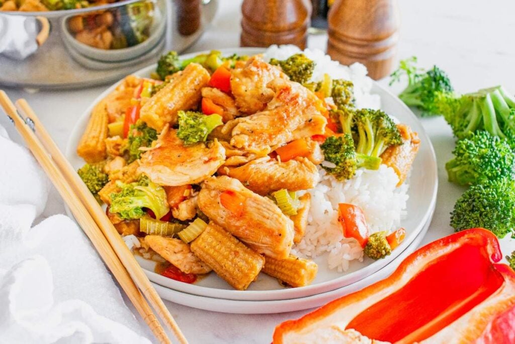 A plate of Hunan chicken stir-fried with broccoli, baby corn, and red bell peppers served on a bed of white rice. Chopsticks rest beside the plate, and fresh vegetables are visible in the background.