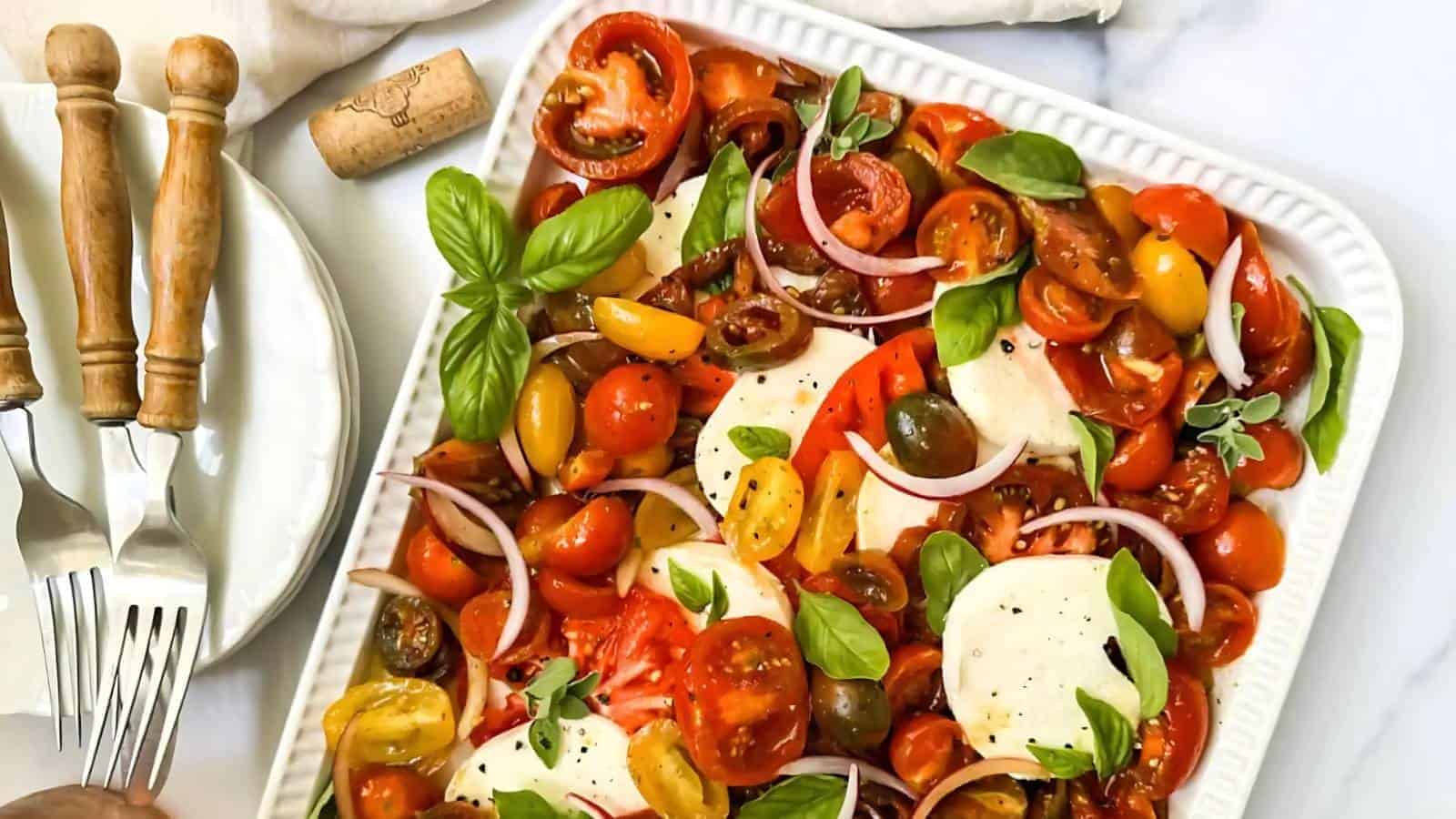 A platter of colorful cherry tomatoes, sliced mozzarella, red onion, and basil leaves. Four forks are placed beside the dish on a white surface.
