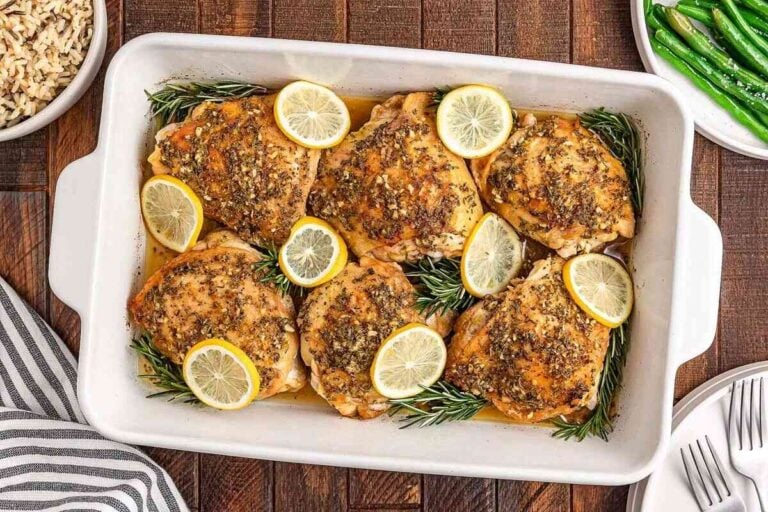Lemon rosemary chicken thighs baked to perfection with lemon slices and fresh rosemary, served in a white dish alongside tender green beans and fluffy rice.