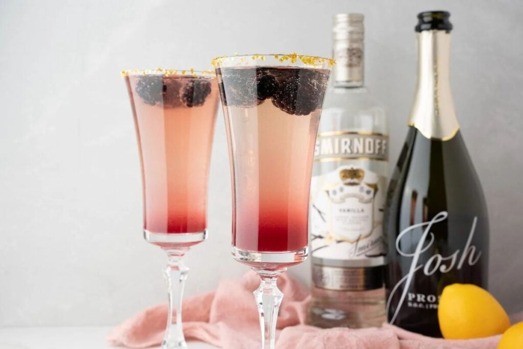 Two berry cocktails in flutes, made with Meyer lemon blackberry syrup, sit beside vodka and prosecco bottles, a lemon, and pink cloth—perfect as a festive Lemon Berry Prosecco Cocktail for your Christmas or New Year’s Eve celebration.