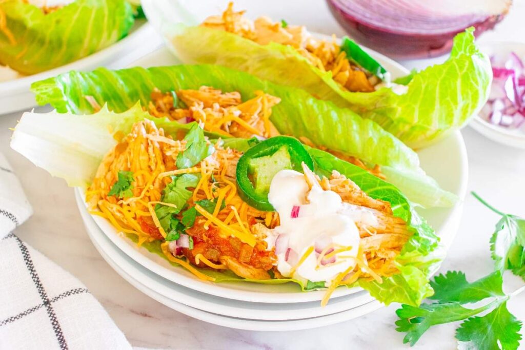 A plate of Instant Pot chicken taco lettuce wraps filled with shredded chicken, grated cheese, sliced jalapeños, and topped with sour cream and cilantro.