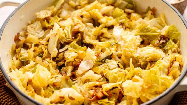 Close-up shot of pan-fried cabbage with browned edges, mixed with small pieces of bacon, in a white enamel cast iron pan.