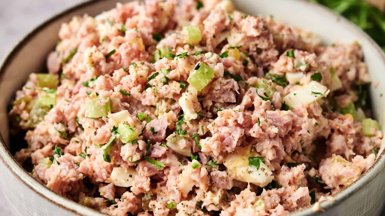 A bowl filled with ham salad, containing chopped ham, celery, hard-boiled eggs, and herbs, mixed with mayonnaise.