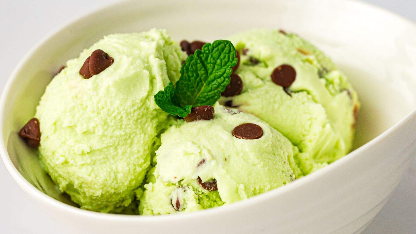 Three scoops of mint chocolate chip ice cream with chocolate chips, garnished with a sprig of mint, in a white bowl.