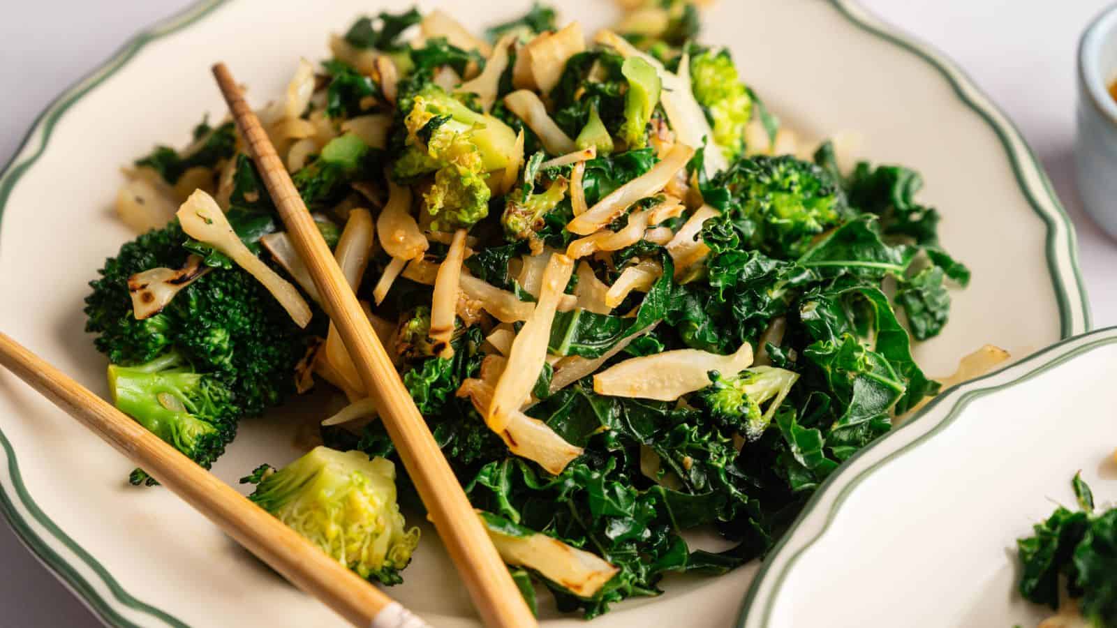 A plate of stir-fried kale, broccoli, and sliced onions, served with a pair of chopsticks on the side.