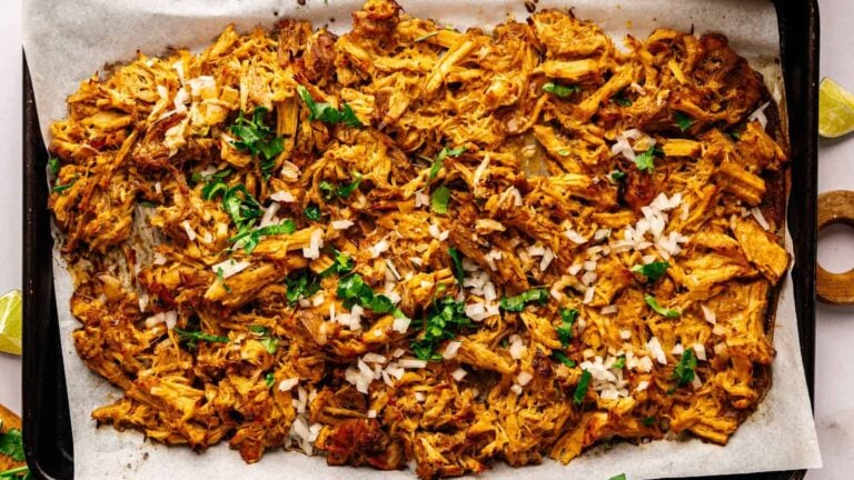 Shredded, seasoned meat spread on a parchment-lined baking sheet, topped with chopped onions and fresh cilantro.