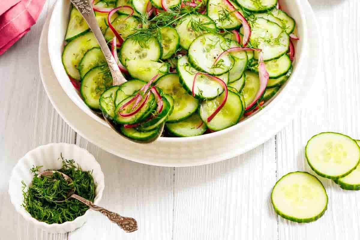 A bowl of cucumber salad with sliced red onions and fresh dill, next to a small dish of chopped dill and cucumber slices on a white wooden surface.