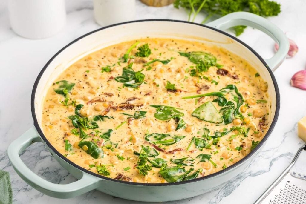  A bowl of creamy Tuscan marry me chickpeas and spinach with herbs and spices simmers in a light blue pot on a marble countertop.