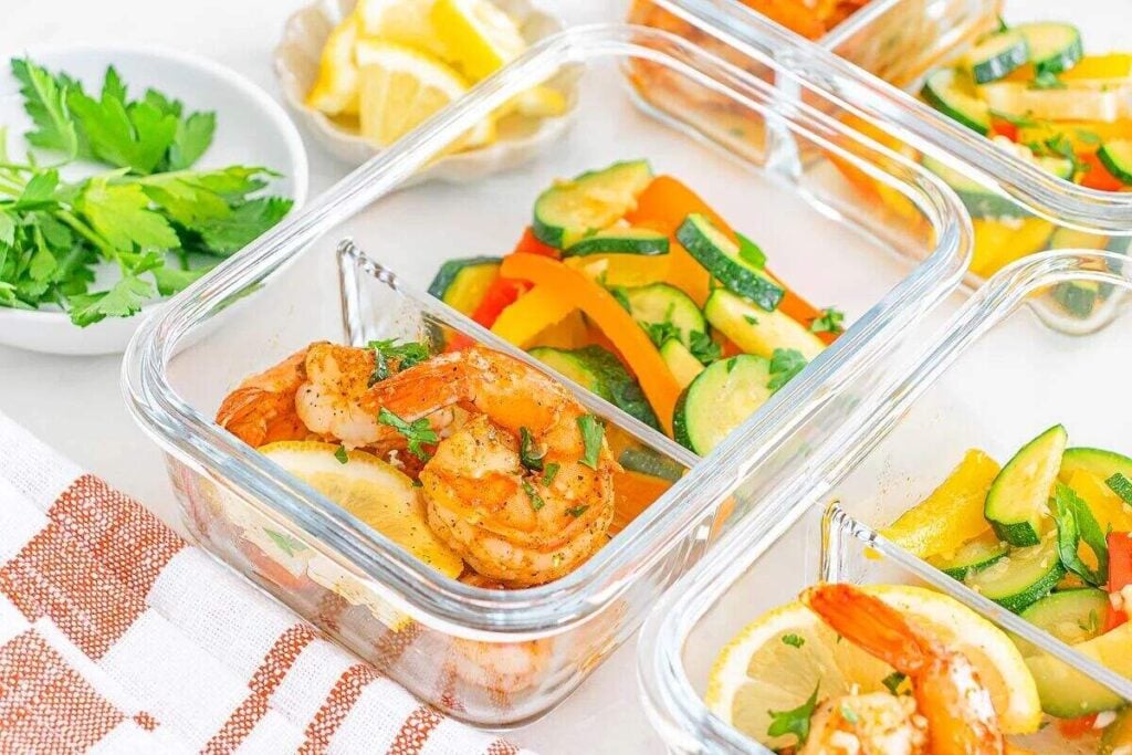 Glass meal prep container with Meal Prep Cajun Shrimp and Veggies, cooked shrimp, lemon slices, and sautéed vegetables next to herbs and lemon wedges.
