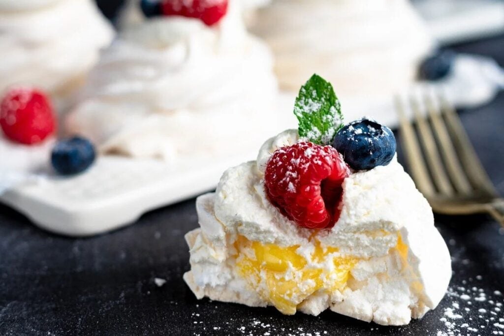 Mini pavlovas with lemon curd, topped with raspberries, blueberries, fresh mint, and dusted with powdered sugar on a dark surface, create an irresistibly elegant dessert with fresh berries.