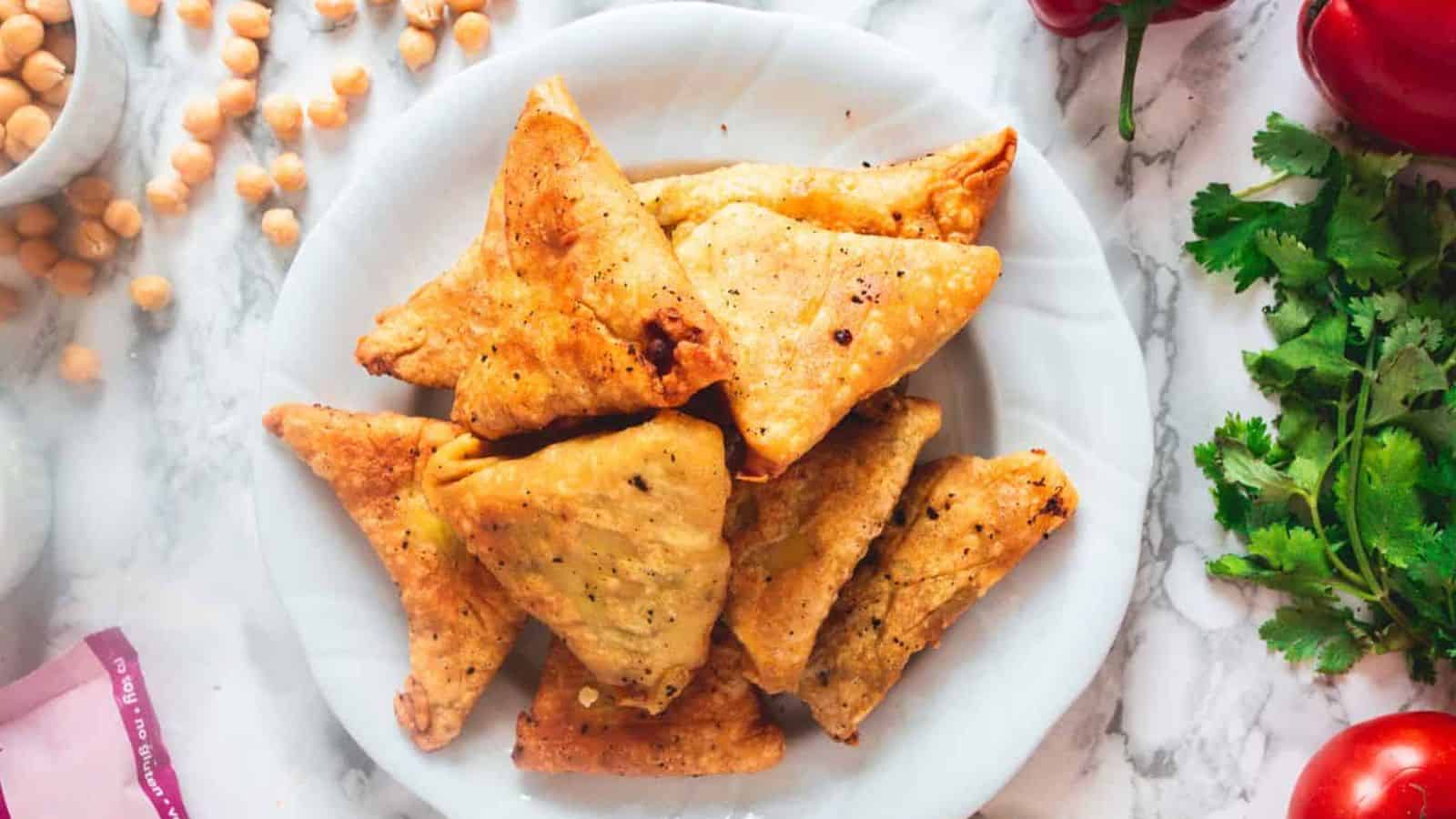 A plate of golden-brown triangular samosas is placed on a marble surface. Surrounding the plate are scattered chickpeas, fresh cilantro, a red pepper, and a cutting board corner. The samosas appear crispy and seasoned.