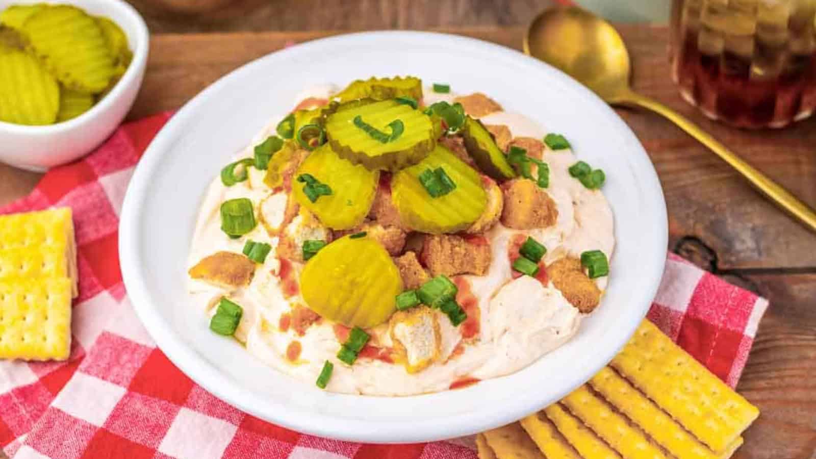 A plate of creamy dip topped with pickles, green onions, and croutons, served with crackers on the side.