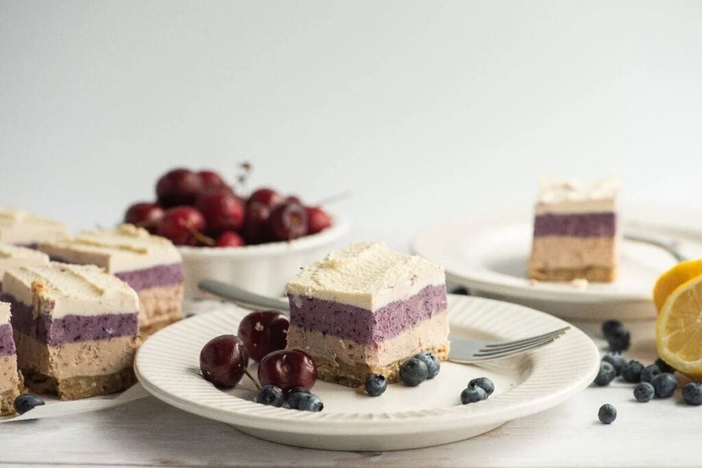 A slice of no bake vegan cheesecake made with raw cashews on a plate with blueberries and cherries, next to other dessert slices.