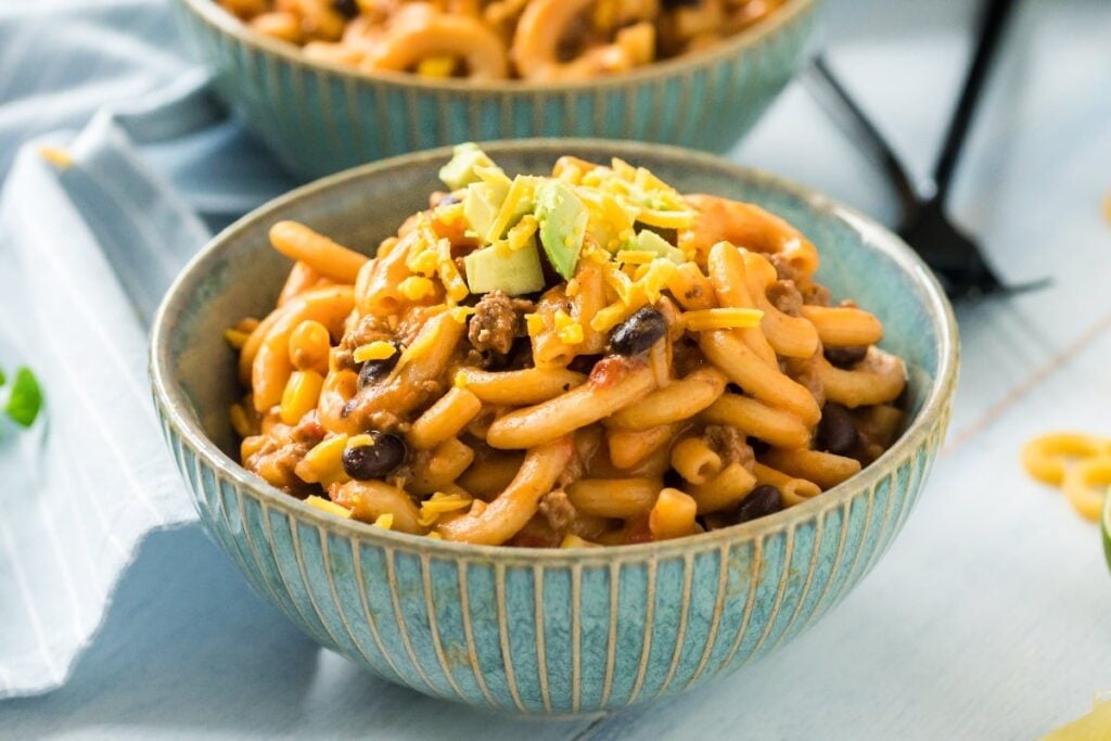 A bowl of easy taco pasta featuring cheesy macaroni, black beans, ground meat, and diced avocado sits invitingly on a light blue table.