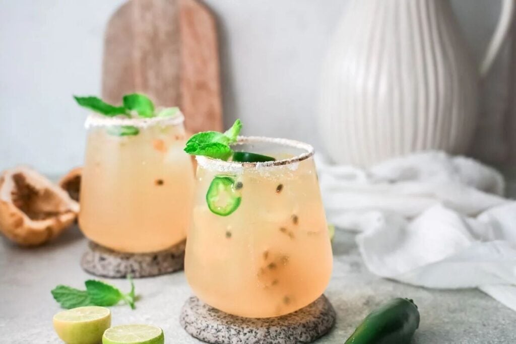 Two cocktails with lime, mint, and jalapeño slices in glasses on stone coasters, these drinks offer a rustic twist—perfect for those seeking an easy margarita recipe with vibrant passion fruit and lime notes.