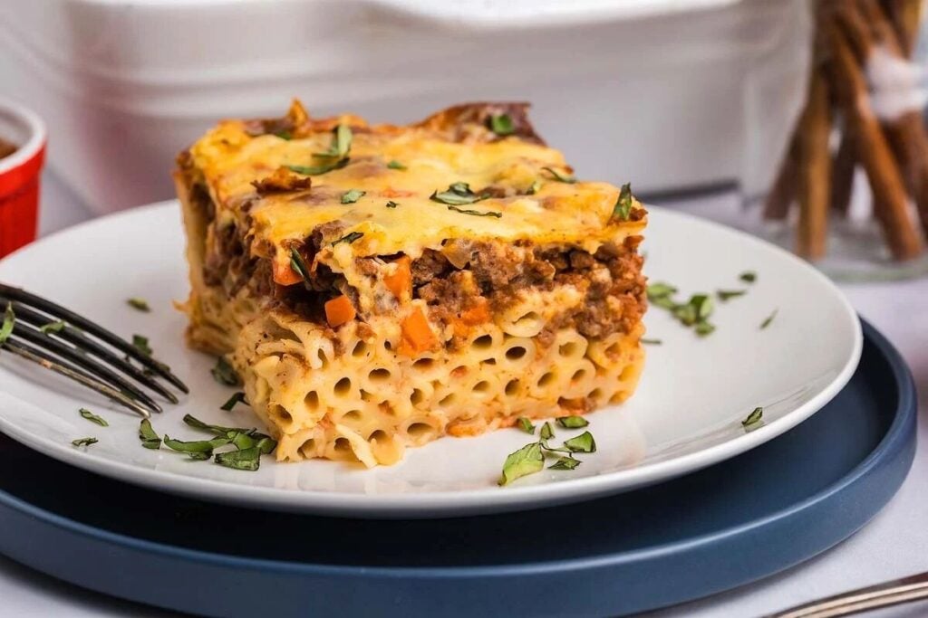 A slice of pastitsio, the classic baked pasta dish featuring ground meat, cheese, and aromatic herbs, rests invitingly on a white plate with a fork nearby.