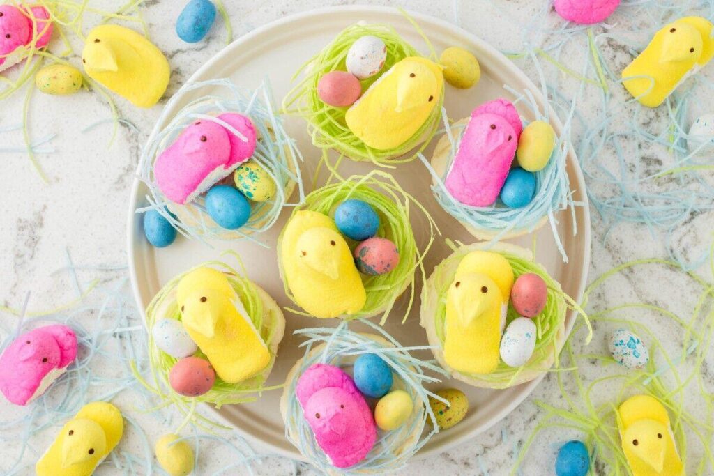 A plate with yellow and pink marshmallow chicks in nests made of green and blue shredded paper, surrounded by colorful candy eggs.