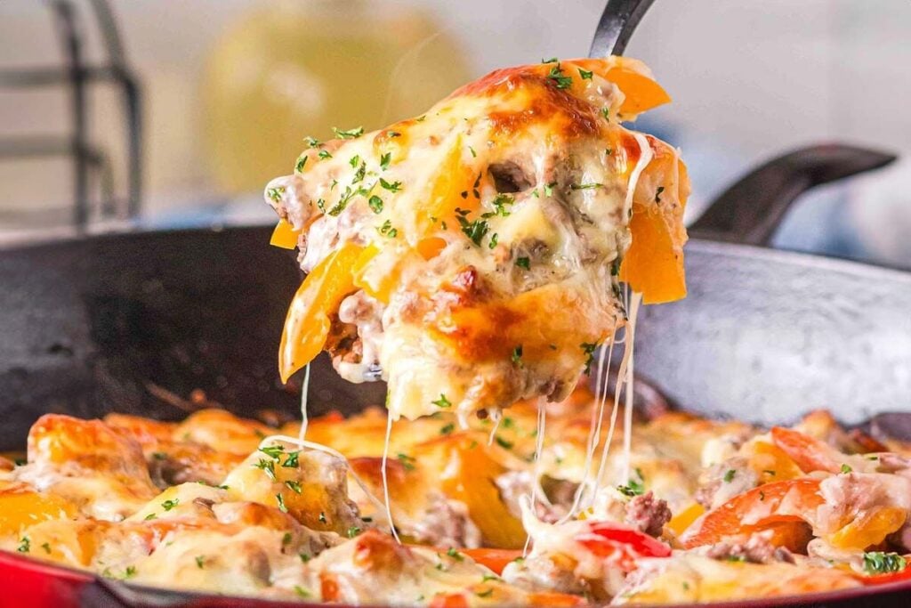 A close-up of a Philly cheesesteak casserole with melted cheese, bell peppers, and ground meat being lifted from a skillet with a serving spatula.