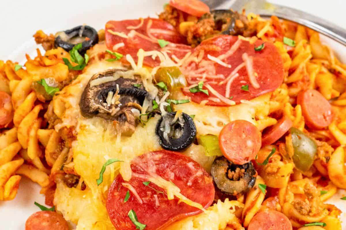 A plate of baked pasta topped with melted cheese, pepperoni slices, black olives, mushrooms, green peppers, and shredded herbs, with a fork on the side.