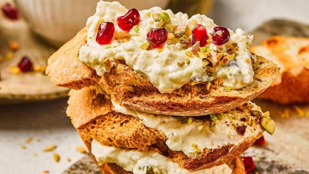 Toasted bread slices topped with creamy cheese, pomegranate seeds, and chopped pistachios.