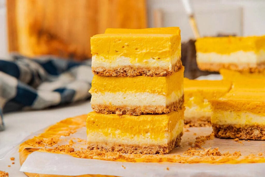 Three stacked pumpkin cheesecake bars with a crumbly crust rest on parchment paper, surrounded by even more delicious pumpkin cheesecake bars.