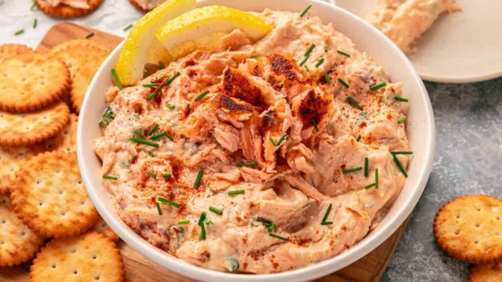 Creamy salmon dip garnished with chives and lemon slices, served with round crackers on the side.