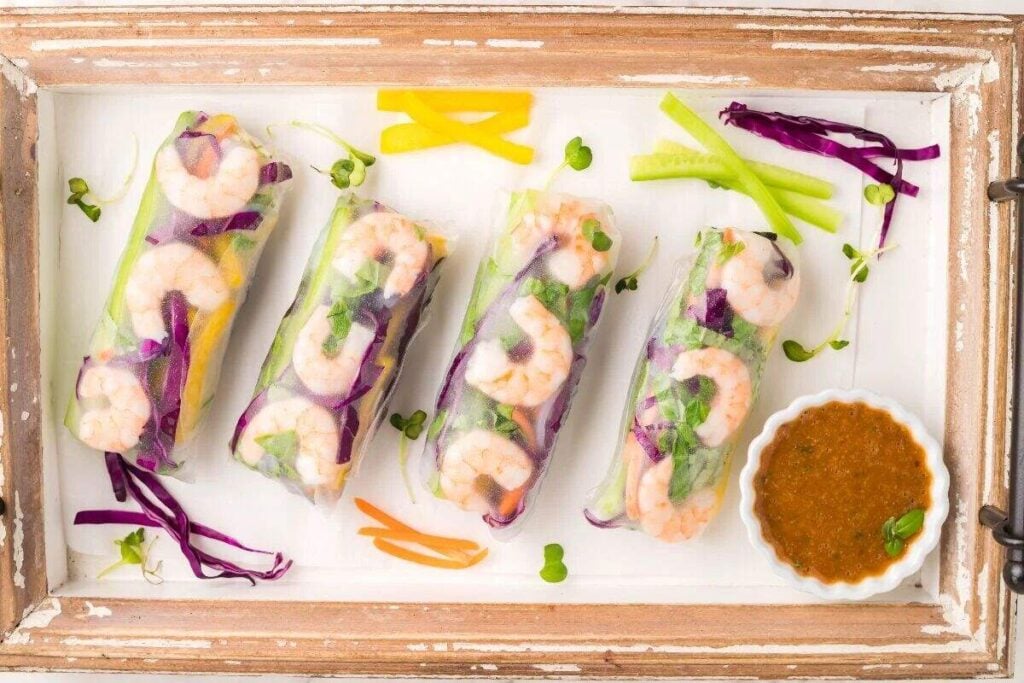 Four shrimp spring rolls with dipping sauce for spring rolls and sliced vegetables on a white tray with wooden edges.
