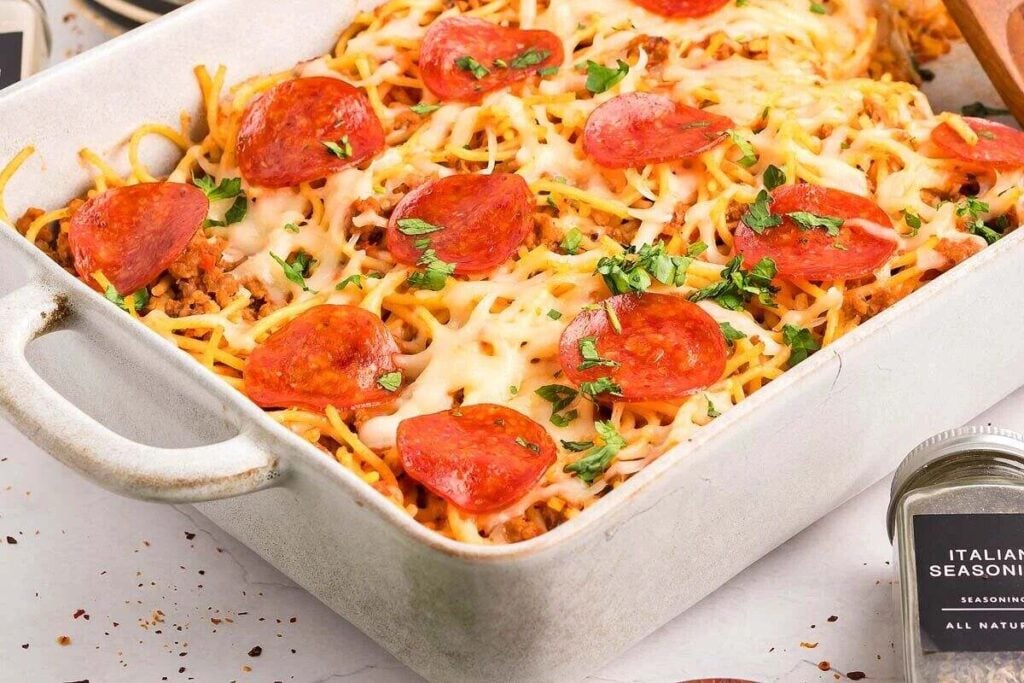 Baked spaghetti pizza casserole topped with melted cheese, pepperoni slices, and chopped parsley in a white dish.