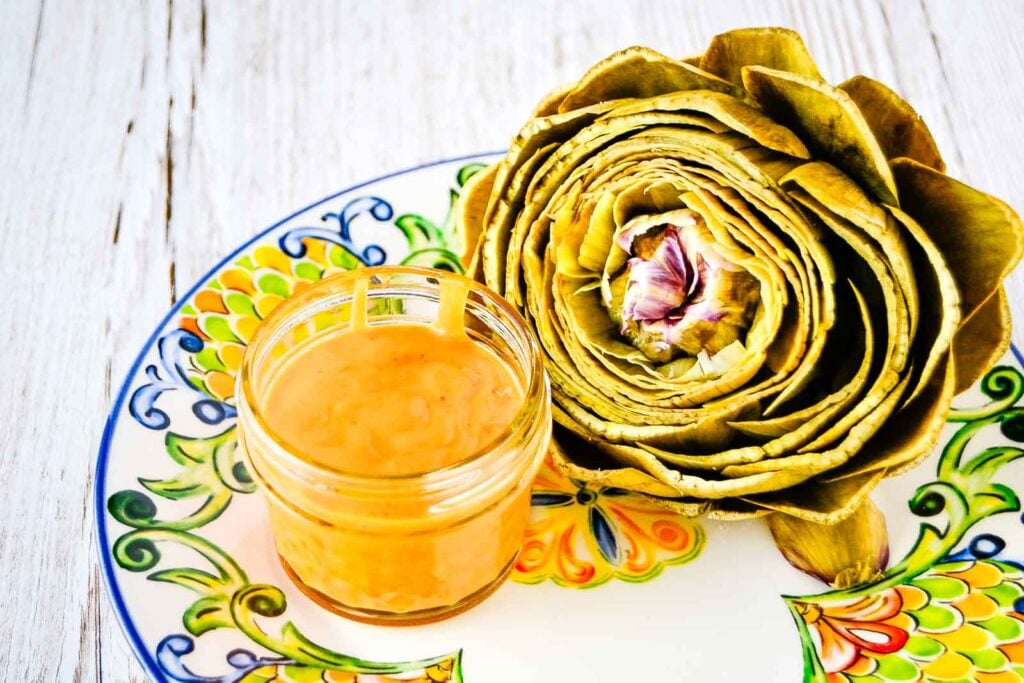 A steamed artichoke on a white plate with sweet and spicy sauce.