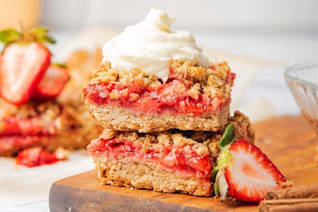 Two strawberry oatmeal bars are stacked on a wooden board, topped with whipped cream, with fresh strawberries and cinnamon sticks in the background.