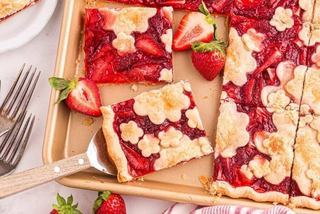 A baking tray holds a delightful strawberry slab pie, its square servings topped with whimsical flower-shaped crusts and surrounded by fresh strawberries.