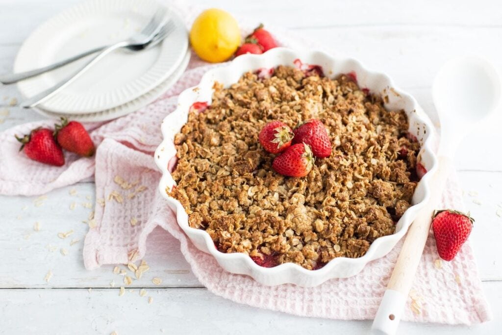 Strawberry Crisp in a white dish, topped with fresh strawberries, sits beside plates and utensils on a pink cloth.