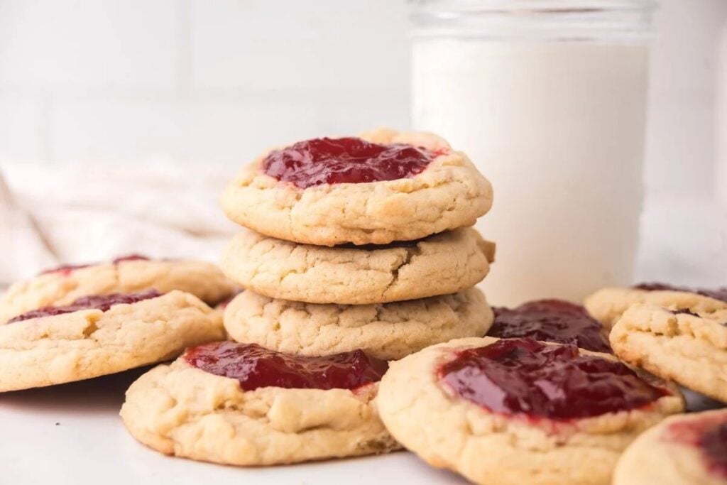 A stack of Strawberry Thumbprint Cookies with red jam centers sits next to a glass of milk on a white surface.