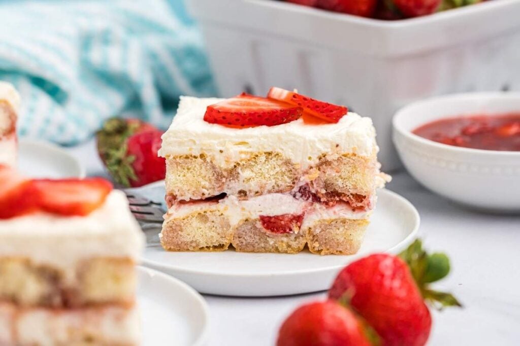 A luscious slice of strawberry tiramisu graces a white plate, adorned with fresh sliced strawberries.