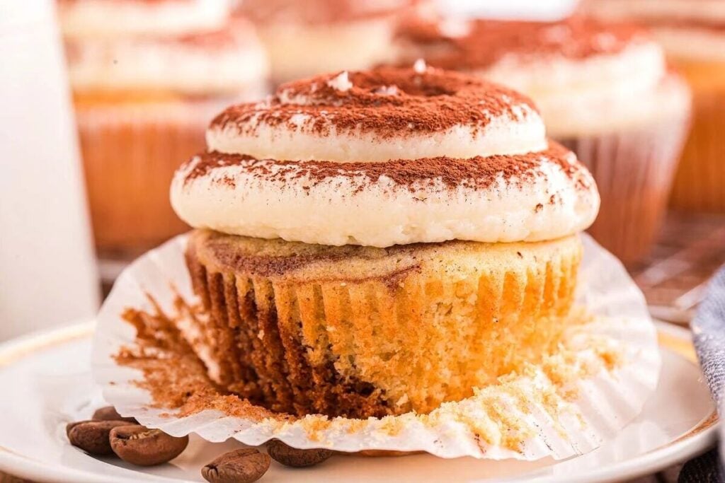 A close-up of delectable tiramisu cupcakes reveals their cream frosting dusted with cocoa powder, each partially unwrapped on a plate adorned with coffee beans.