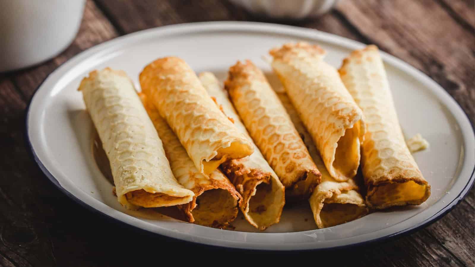 Six rolled Norwegian krumkake cookies on a white plate, set on a rustic wooden table.