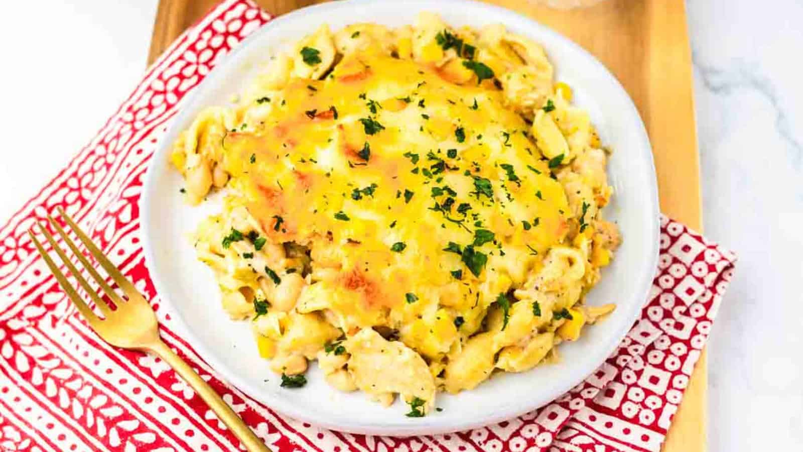 A plate of cheesy pasta casserole garnished with chopped parsley, on a red-patterned napkin with a gold fork.