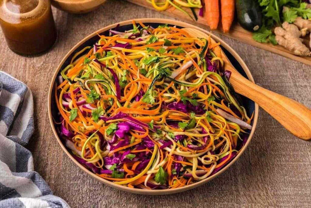 Bowl of vibrant Asian Zoodle Salad with carrots, cabbage, and fresh herbs on a wooden table.