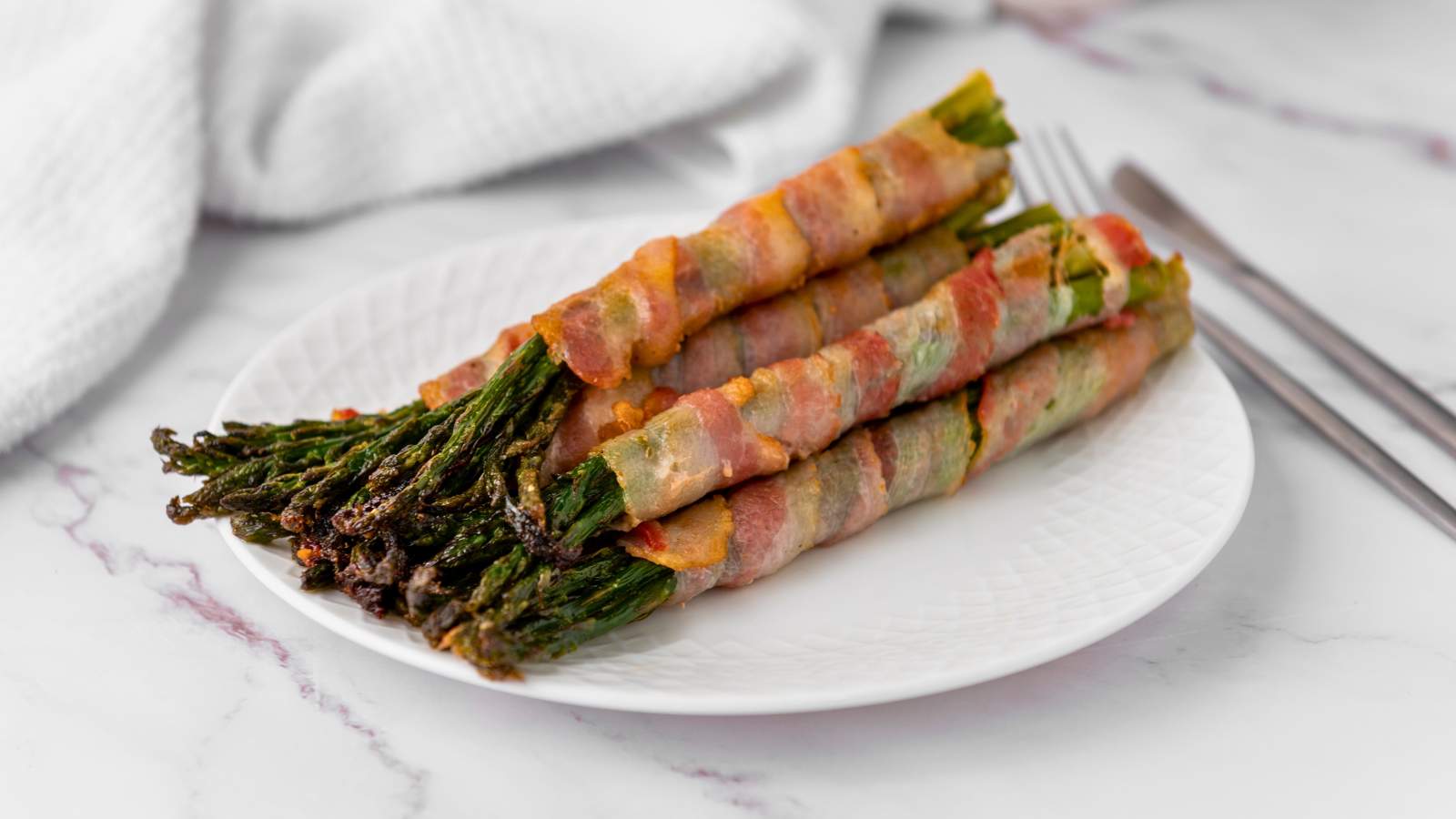 A white plate with bacon wrapped asparagus placed on a marble surface with a fork and knife nearby.