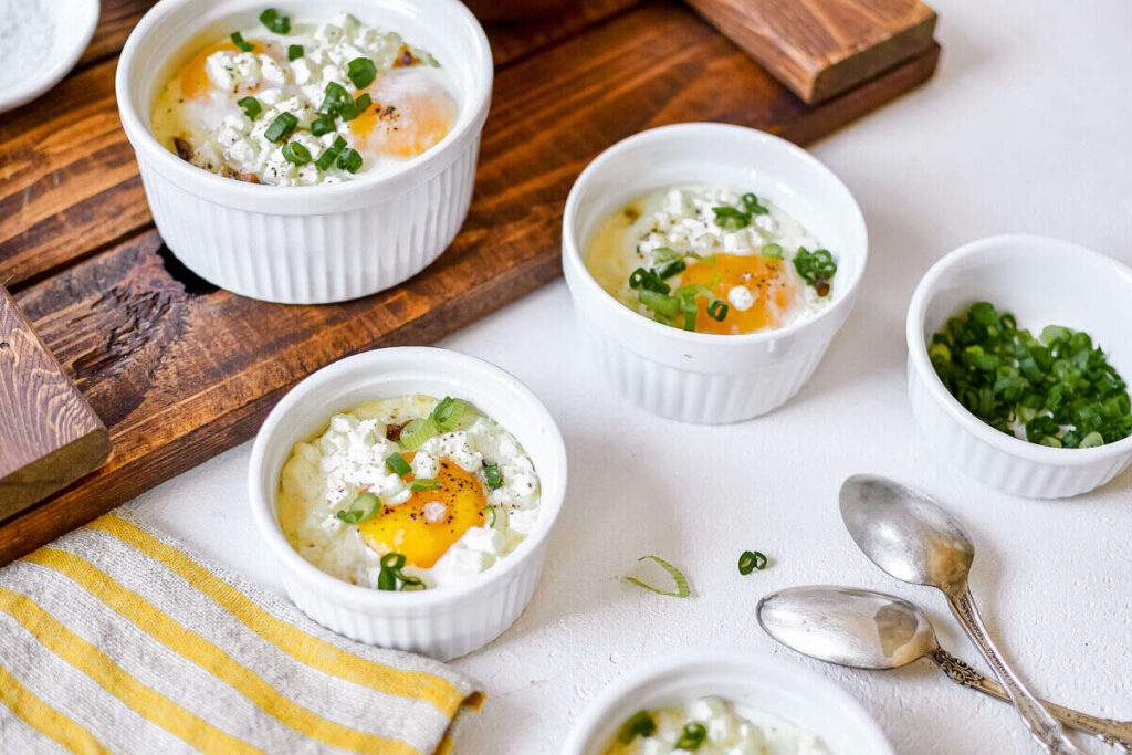 Baked eggs with cheese and green onions in white ramekins, with spoons and a bowl of chopped green onions nearby.