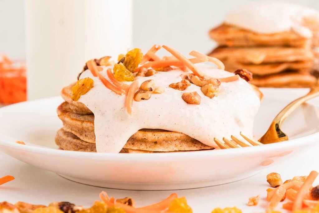 A stack of carrot cake pancakes topped with cream, shredded carrot, raisins, and walnuts sits enticingly on a white plate with a fork.