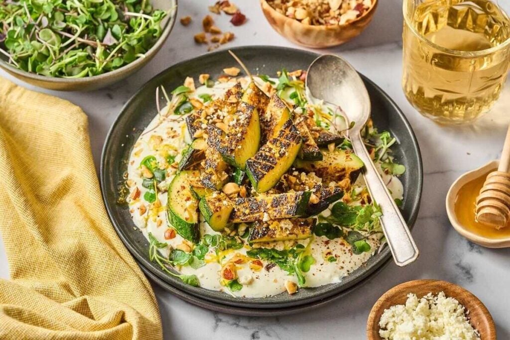 Grilled zucchini on a plate with a creamy cottage cheese sauce, topped with nuts and greens. 