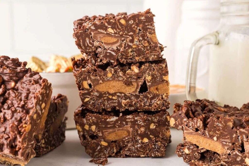A stack of chocolate peanut butter crunch bars sits on a table, with a glass jar in the background.