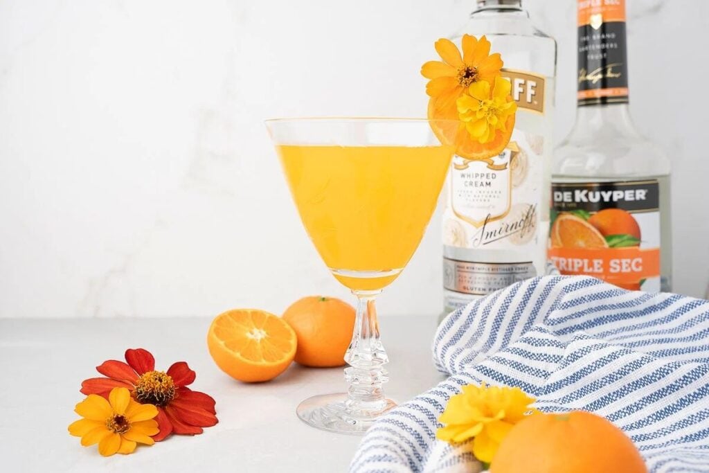 A Clementine Martini, made with whipped cream flavoured vodka and triple sec, sits in a glass with an orange flower garnish, surrounded by fresh mandarins and bottles of liquor—a perfect summer martini recipe.