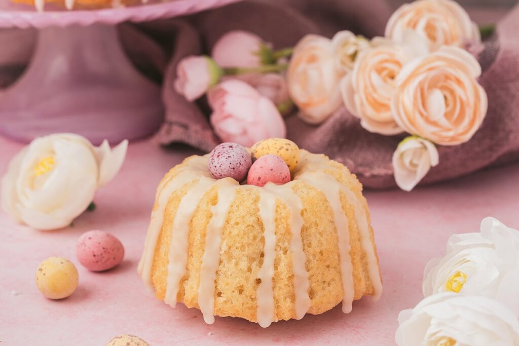 A vanilla drizzle mini cake decorated with candy eggs and surrounded by pastel-colored flowers, now enhanced as an Easter dessert.