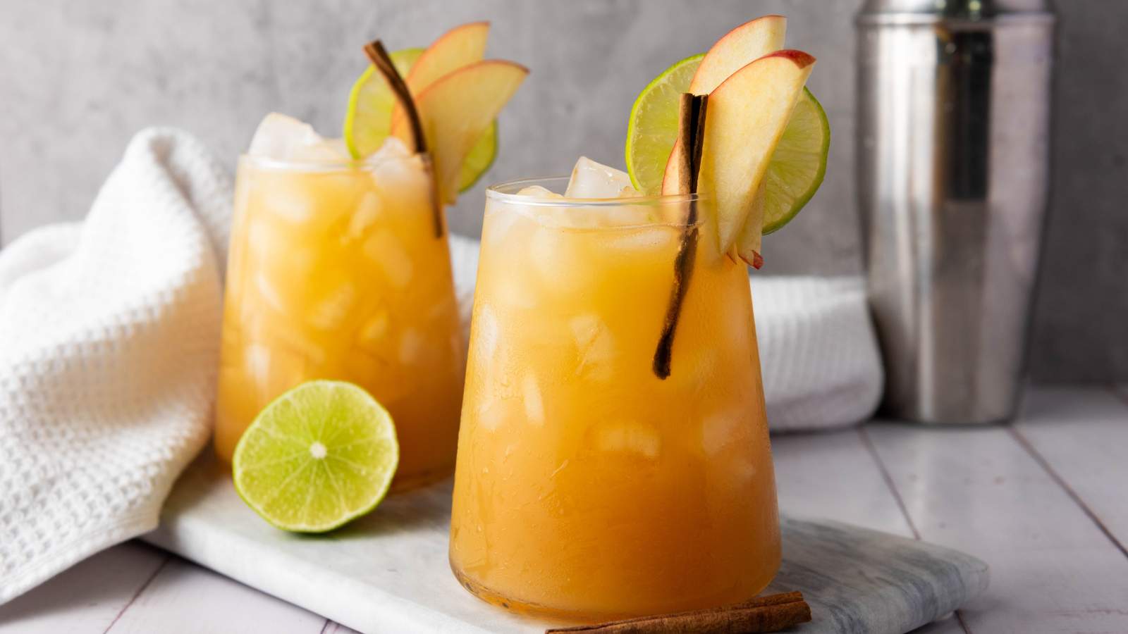 Two glasses of orange-colored iced drinks garnished with apple slices, lime wheels, and cinnamon sticks on a marble board, with a lime half, cinnamon stick, towel, and cocktail shaker nearby.
